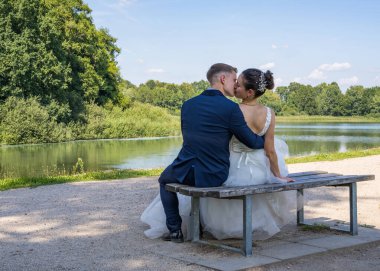 Genç ve güzel gelin ve damat suyun önündeki bankta oturuyorlar. Evli bir çiftten şefkatli bir öpücük. Arka plan.