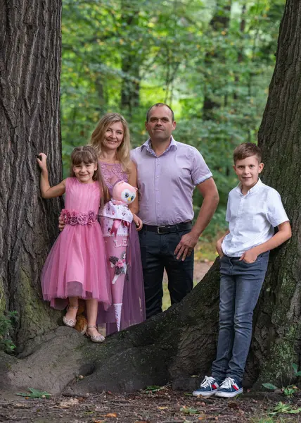 Portrait of a happy family. A laughing father and mother, a teenage son and a first-grader daughter stand near a large tree in the park. Mom and girl are dressed in beautiful pink dresses. A happy family