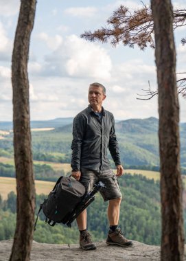 Fotoğrafçı dağların tepesinde duruyor, elinde bir sırt çantası ve kameraya bağlı bir tripodla. Sakson İsviçre, Almanya
