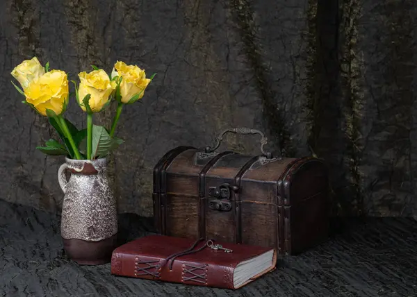 Still life with roses. Three yellow roses in a vase on a background of dark fabric, an old wooden chest, a notebook in a leather cover