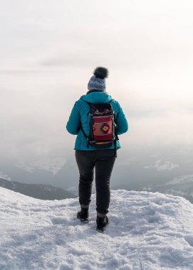 Sırtında süs olan bir sırt çantasıyla bir kadın bir dağın kenarında duruyor ve kar kaplı dağlarda yürürken manzaranın tadını çıkarıyor. Sniezka Karkonosze 