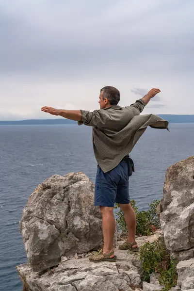 Gezinin tadını çıkaran orta yaşlı bir adam. Uçurumun kenarında duruyor ve deniz manzarasına bakıyor. O, kollarını yanlara açtı. Gömlek rüzgarda dalgalanıyor..