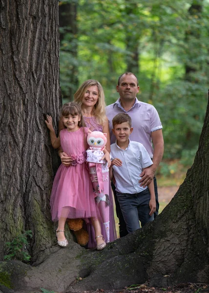 Mutlu bir ailenin portresi. Gülen bir baba ve anne, genç bir oğlan ve birinci sınıf bir kız çocuğu parkta büyük bir ağacın yanında duruyor. Anne ve kız güzel pembe elbiseler giydiler. Mutlu bir aile.