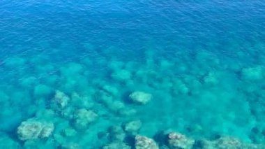 Mavi şeffaf deniz, dipte kayalar görünüyor.