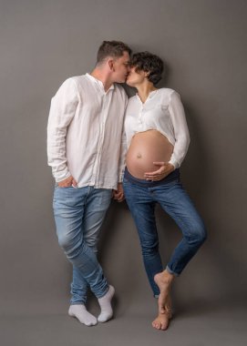 Çıplak göbekli hamile bir kadın duvara yaslanır ve kocasını öper. İkisi de beyaz gömlek ve kot pantolon giyiyor.