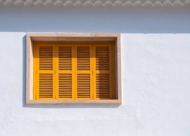 Beyaz bir duvarda sarı ahşap panjurlar olan küçük bir pencere..