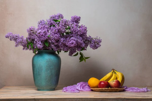 A large bouquet of lilacs in a vase on a light gray background, next to them on a plate are bananas, two red apples, and an orange. Still life, plenty of space for text