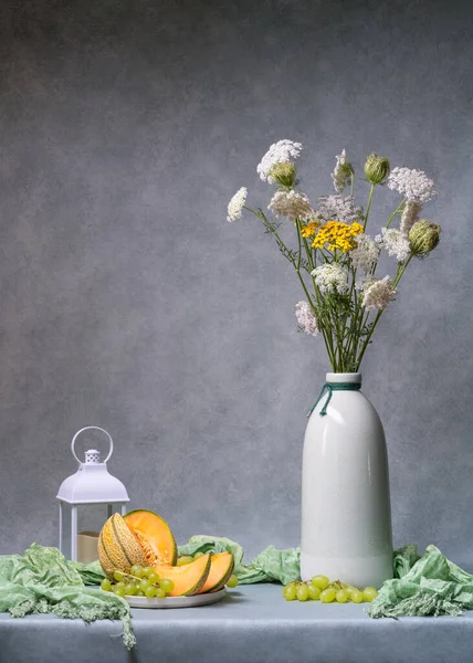 Still life with flowers. White wildflowers in a light tall vase on a light gray background, next to it lies a cut orange melon, a bunch of white grapes, and a lamp. Plenty of space for text