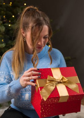 Sarı saçlı, mavi kazaklı genç bir kadın üzerinde altın bir fiyonk olan kırmızı hediye kutusunu açıyor. İçeriden aydınlatılmış açık hediye kutusu.