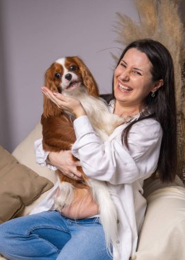 Beyaz gömlekli, mavi kotlu, göbekli güzel, genç ve hamile bir kadın evdeki kanepede oturuyor. Güzel beyaz ve kahverengi bir cocker köpeğini kollarında tutuyor.