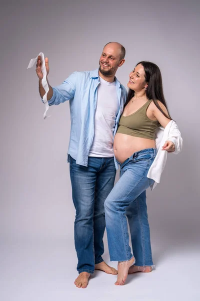 A pregnant woman in a white shirt, blue jeans with her belly exposed and her husband are holding a photo of their unborn newborn baby. Pregnancy, motherhood