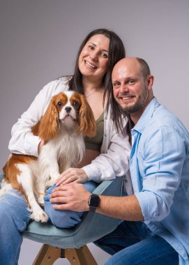 Çıplak göbekli hamile bir kadın mavi bir koltuğa oturur, bir cocker spaniel 'i kucağına alır ve nazikçe kocasına sarılır.