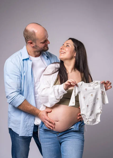 A pregnant woman in a white shirt, blue jeans with a bare belly and her husband are holding clothes on the baby.On a white background.Pregnancy, motherhood