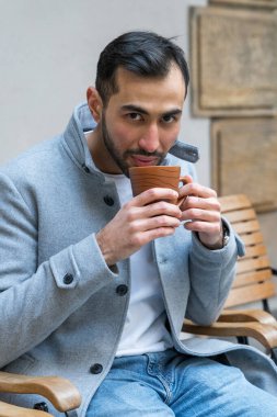 Asyalı yakışıklı, düzgün sakallı, açık renk ceketli genç bir adam bankta oturuyor ve elinde bir fincan çay tutuyor..