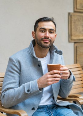 Asyalı yakışıklı, düzgün sakallı, açık renk ceketli genç bir adam bankta oturuyor ve elinde bir fincan çay tutuyor..