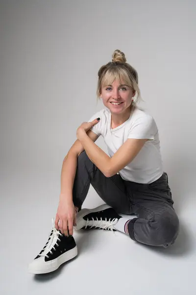 Full length photo of an adorable blonde woman sitting on the floor, wearing jeans, white t-shirt and sneakers, isolated on a white background. Smiles cheerfully and looks at the camera