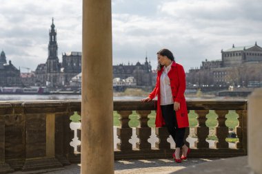 Koyu saçlı, kırmızı paltolu güzel bir bayan turist, Almanya 'nın eski kasabası Dresden' in köprüsünden güzel bir manzaranın tadını çıkarıyor.