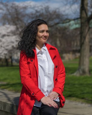 Kırmızı ceketli ve beyaz bluzlu şık bir kadının romantik portresi. Pozitif ruh hali. Bir bayan şehir parkında yürüyor, korkuluğa oturuyor ve güneşli bir günün tadını çıkarıyor.