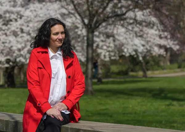 Romantic portrait of a stylish woman in a red coat and white blouse. Positive mood. A lady walks through a city park, sits on the parapet and enjoys a sunny day