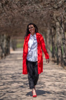 Havalı, kendine güvenen bir kadın. Koyu renk saçlı, kırmızı ceketli, beyaz bluzlu ve kırmızı topuklu ayakkabılı bir model parktaki bir ara sokakta neşeyle uçuyor. Bahar modası eğilimi