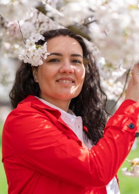 Kırmızı ceketli ve beyaz bluzlu şık bir kadının romantik görüntüsü. Pozitif ruh hali. Tatlı bir kız nazikçe beyaz bir sakura dalı tutuyor ve kameraya bakıp gülümsüyor.