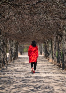  Dışarıdaki şık bir kadının portresi. Koyu renk saçlı, kırmızı yağmurluklu ve kırmızı topuklu ayakkabılı bir model parktaki ağaçların arasında bir ara sokakta yürüyor. Arka plan