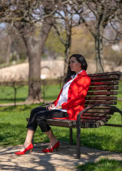 Romantic portrait of a stylish, beautiful woman in a red coat and white blouse, red shoes. Positive mood. A lady walks through a city park, sits on a bench and enjoys a sunny day.