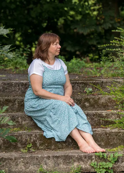 A middle-aged woman sits on old stone steps in an abandoned park. Dressed in a long dress and without shoes, barefoot. Looking thoughtfully into the distance