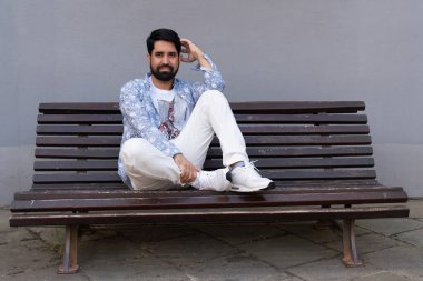 Modern, şık, siyah sakallı, açık renk elbiseli Pakistanlı bir adam, ahşap bir bankta oturuyor.