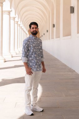 Modern, şık, siyah sakallı ve açık renk elbiseli Pakistanlı bir adam beyaz sütunların arasında duruyor.
