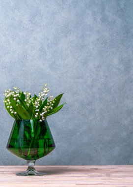 White flowers, lilies of the valley in a green transparent vase, a glass, next to it lies a green scarf and three green balls. Gray background. Plenty of space for text clipart