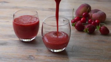   Bir kadının eli, ahşap masaya çilekli meyve suyu döker. İki bardak çilekli smoothie. Yakınlarda meyve var. Yaz içeceği. Yakın plan. 
