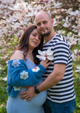 Bir erkek, hamile karısına şefkatle sarılır. Kadın mavi bir elbise giyiyor. Manolya çiçeklerinin yanında bir çift erkek ve hamile kadın.