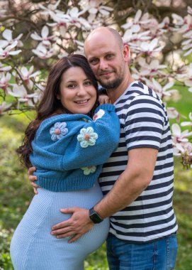 Bir erkek, hamile karısına şefkatle sarılır. Kadın mavi bir elbise giyiyor. Manolya çiçeklerinin yanında bir çift erkek ve hamile kadın. 