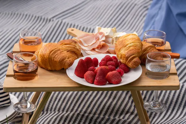 A small table served with picnic food stands on a green clearing in a city park. Strawberries, croissants, cheese, snacks, wine Outdoor picnic, various snacks,