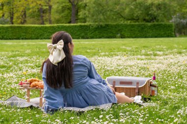 Mutlu kadın hayattan zevk alıyor. Doğada zaman geçiriyor. Parktaki çimlerin üzerinde oturan bir bayan, yanında yemek olan bir piknik masası var. Arkadan bak. Kafasında beyaz bir fiyonk var. Elinde bir bardak tutuyor.