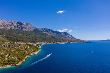 Turistlerle dolu küçük bir tekne, berrak sularda yavaşça yüzüyor. Harika bir kıyı şeridi. Hava fotoğrafçılığı. Dağlar yeşillikle kaplıdır, mavi şeffaf deniz. Makarska Riviera. Hırvatistan 
