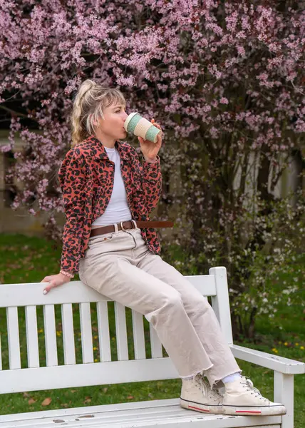 一个漂亮的年轻女子沉着地喝着一杯咖啡的侧视图 一个金发女郎坐在公园的长椅上 享受着生活 在繁忙的生活节奏中稍作停顿 — 图库照片