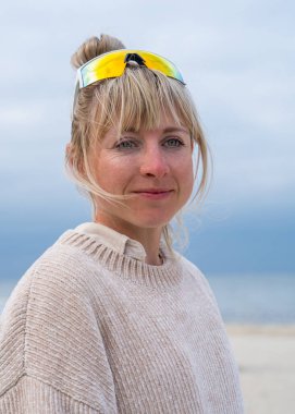 Sarı saçlı, gülümseyen güzel bir kadının portresi. Kafasında güneş gözlüğü var. Hafif giysiler içinde. Denizin arka planında duruyor. 