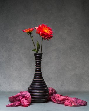 İki kırmızı çiçekle sakin bir hayat. Dahlias, gri arka planda, koyu, ahşap bir vazoda. Güzel minimalist çiçek dekoru. Boşluğu kopyala. 