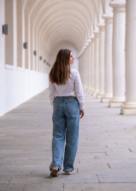 Bir esmerin tam boy dikiz manzarası. Beyaz kolonlar arasında uzun saçlı şık bir genç kadın yürüyor.