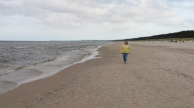 Mutlu orta yaşlı bir kadın deniz boyunca sakin bir şekilde yürür. Dalgalar kumlu kıyıya vuruyor. Baltık Denizi 'nde tatiller, huzur, rahatlama..