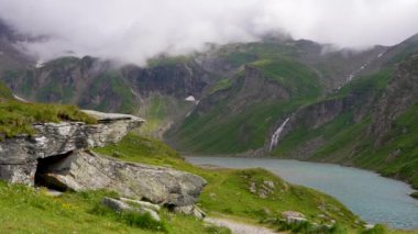 Dağlar yeşil çimlerle kaplıdır. Kalın bulutlar. Şelale aşağı akar. Turkuaz göl. Doğanın güzelliği büyüleyici. Grossglockner yüksek alp yolu. Avusturya 