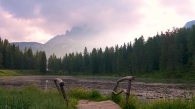 Ünlü Drei Zinnen ya da Tre Cime di Lavaredo dağının üzerinde pembe gün doğumu. Kalın bulutlar yoğun bir dağ örtüsü altında. Ön planda eski ahşap bir köprü. Dolomite Alpleri 'nde sabahın erken saatlerinde. İtalya.