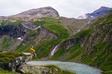 İnsan, dağların etrafını çevreleyen yeşilliklerle çevrili dağ gölüne bakarak dev bir kayanın üzerinde oturur. Şelale uzaktan görülebilir. İnsanoğlu doğanın güzel manzarasının tadını çıkarıyor.