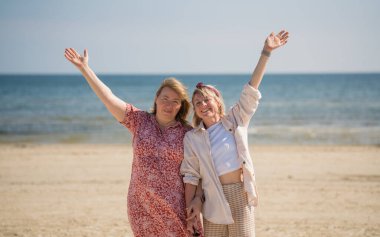 Anne ve yetişkin kız kumsalda elleri havada, sakin denize ve açık gökyüzüne karşı mutlu bir şekilde gülümsüyorlar. Mutlu bir aile. Tatilde. Farklı nesiller