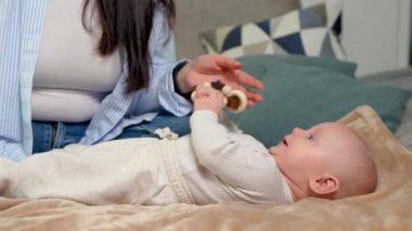 Sevimli yeni doğmuş bebek çıngırakla oynuyor, elinde tutuyor ve yatakta sırtüstü yatarken ona bakıyor. Annem bebeğe yaslanıyor. Çocukların motor becerilerini geliştirmek için oyuncaklar.