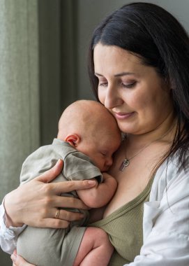Kadın yeni doğmuş oğlunu şefkatle kucaklıyor. Anne bebeğe içtenlikle bakıyor. Bebek rahatça yuvalanmış ve kollarında huzur içinde uyuyor. Anne bakımı ve sevgi.