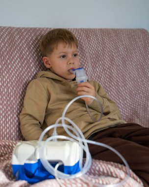 Anaokulu çocuğu evdeki kanepede otururken nefes alıyor. Çocuk soğuk algınlığını tedavi etmek için ilaç cihazının yardımıyla nefes alıyor. Evde tedavi görüyor. Solunum terapisi.