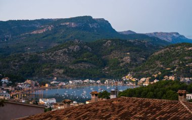 Evening Port de Soller. On seafront lights of cozy cafes and restaurants. Boats and yachts are swaying near the shore. Narrow bay is surrounded by mountains. Evening Mallorca, Spain. clipart